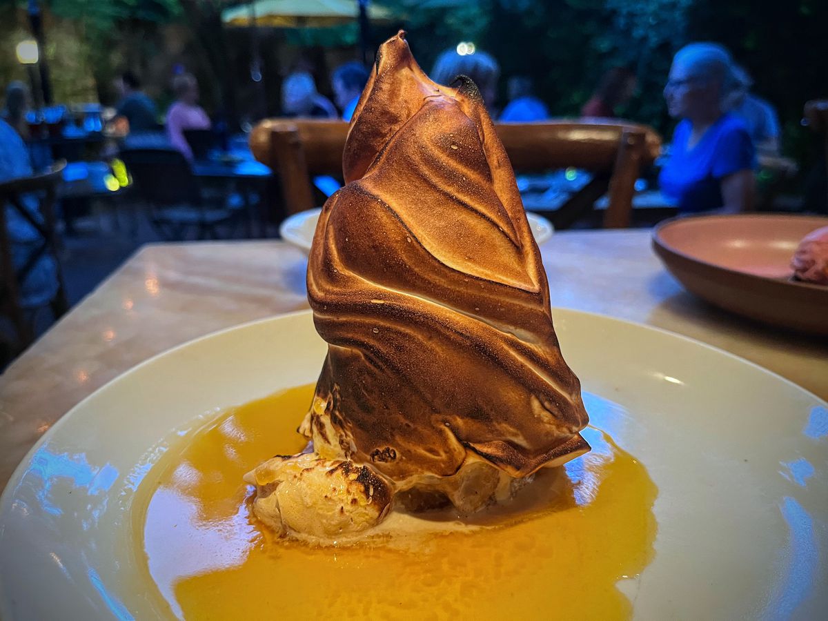 A tall baked Alaska with a tower of toasted meringue sits on a white plate in a pool of yellow sauce.