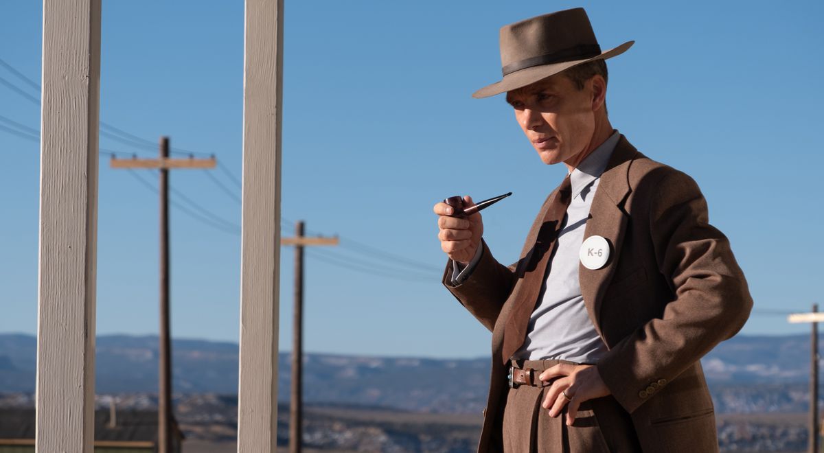 J. Robert Oppenheimer (Cillian Murphy), in brown suit and hat, holds a pipe and stands in a desert near a row of telephone poles in Christopher Nolan’s Oppenheimer