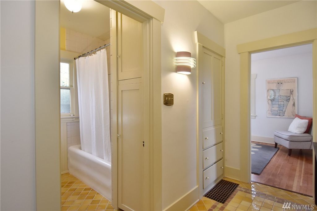 A corner of a tiled room features a built-in pantry. A bathroom is to the left, a room with hardwood floors to the right.