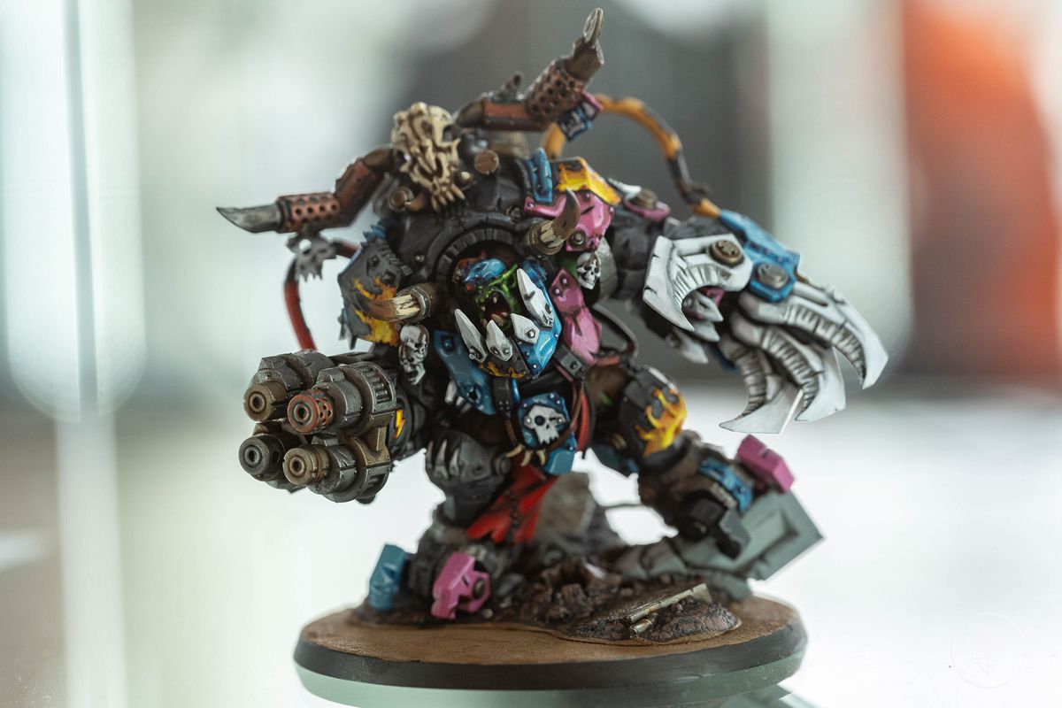 An ork warlord in magenta and light blue — the colors of the trans pride flag. The model is of an ork that has transformed itself mostly into a machine. Its guns are silent, but show wear and age — as if cobbled together from four or five other firearms. Different metallic finishes complete the illusion.
