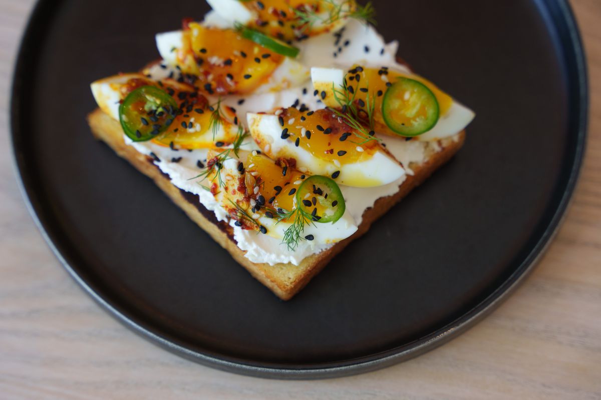 A tartine topped with eggs, sesame seeds, chili oil, and thin slices of jalapeno on a black plate on a wooden table. 
