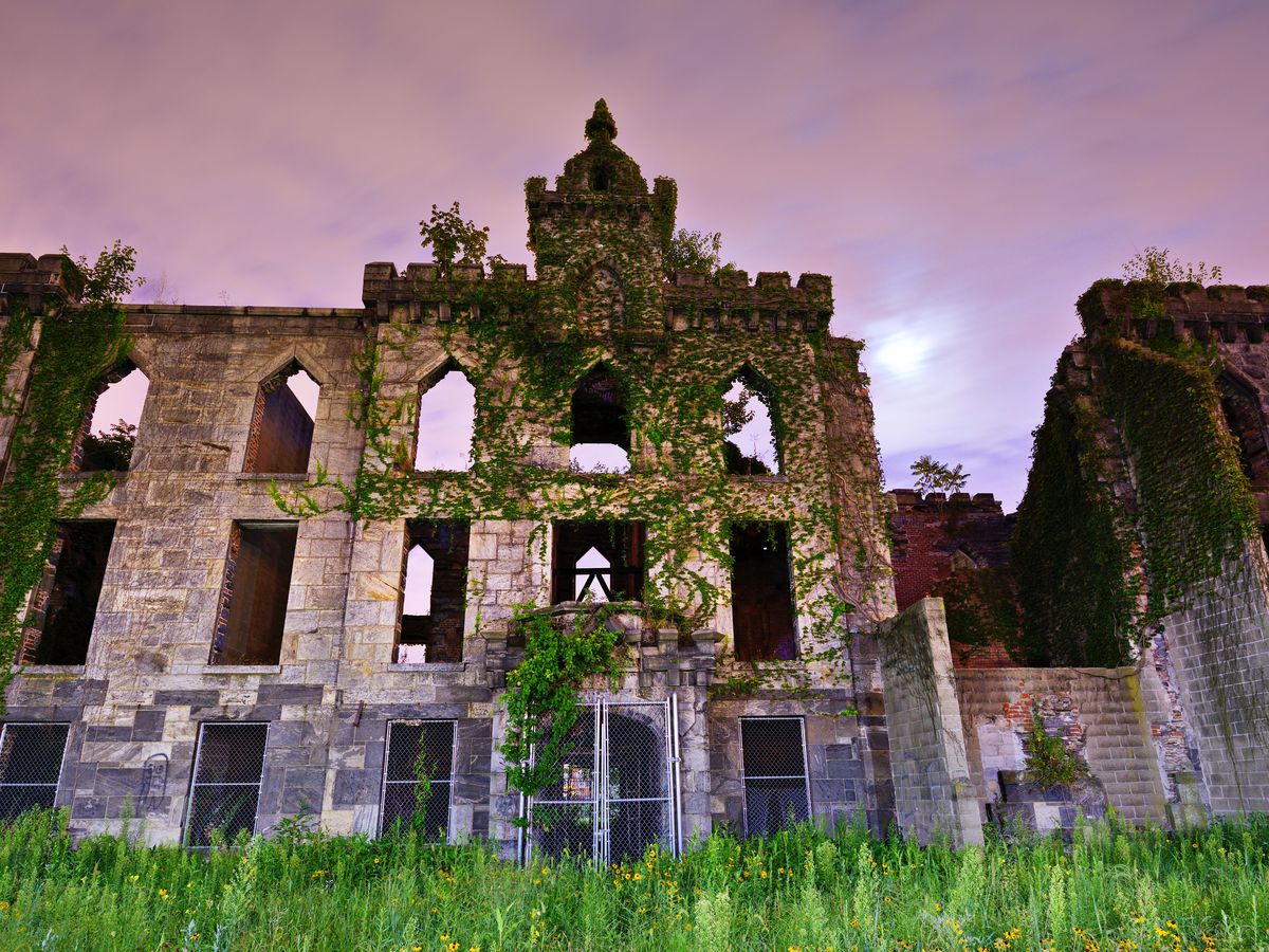 The exterior of the Renwick Smallpox Hospital. The building is abandoned. There are multiple windows and the facade is stone. There is moss growing on some parts of the facade. It is evening and the sky is purple.