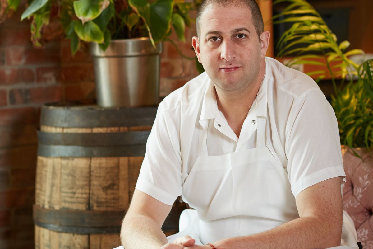 A photo of chef Steve “Nookie” Postal, wearing chef’s whites and sitting in front of a barrel and greenery in a restaurant