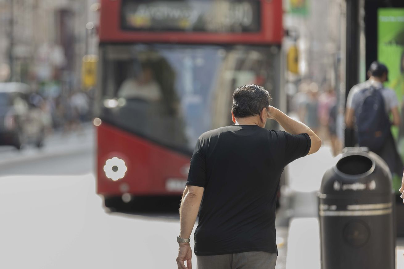 A man touches his face as he walks towards a red-double-decker bus.