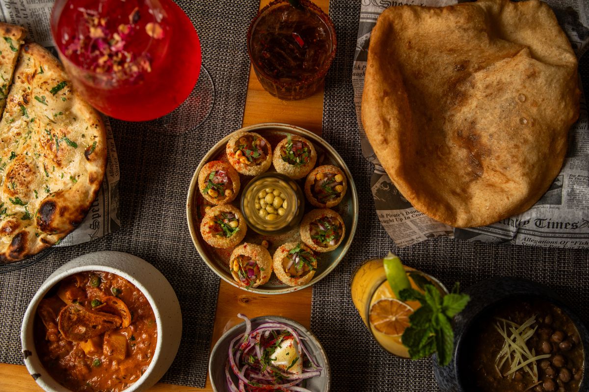 Overhead shot of naan, chole bhature, pani puri, puffed bread, and cocktails.