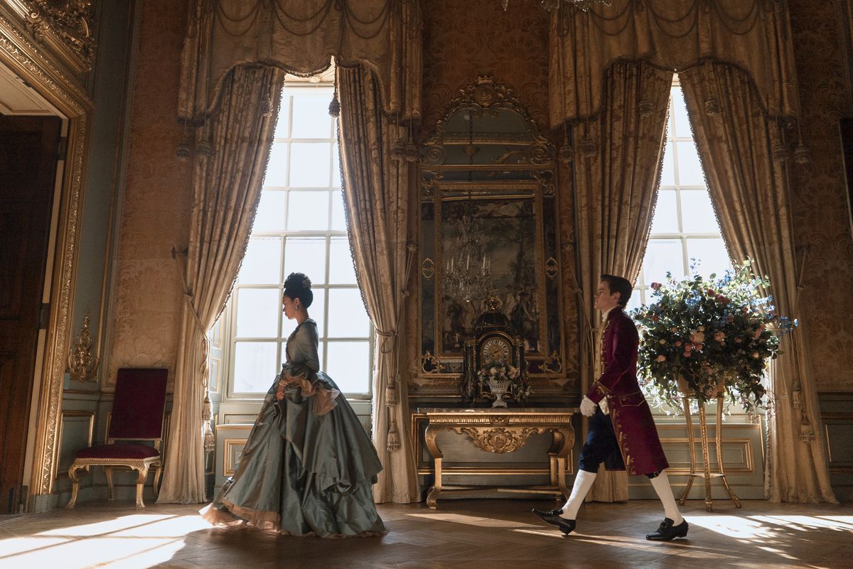 young queen charlotte walks in the fancy halls of the palace, a manservant following her
