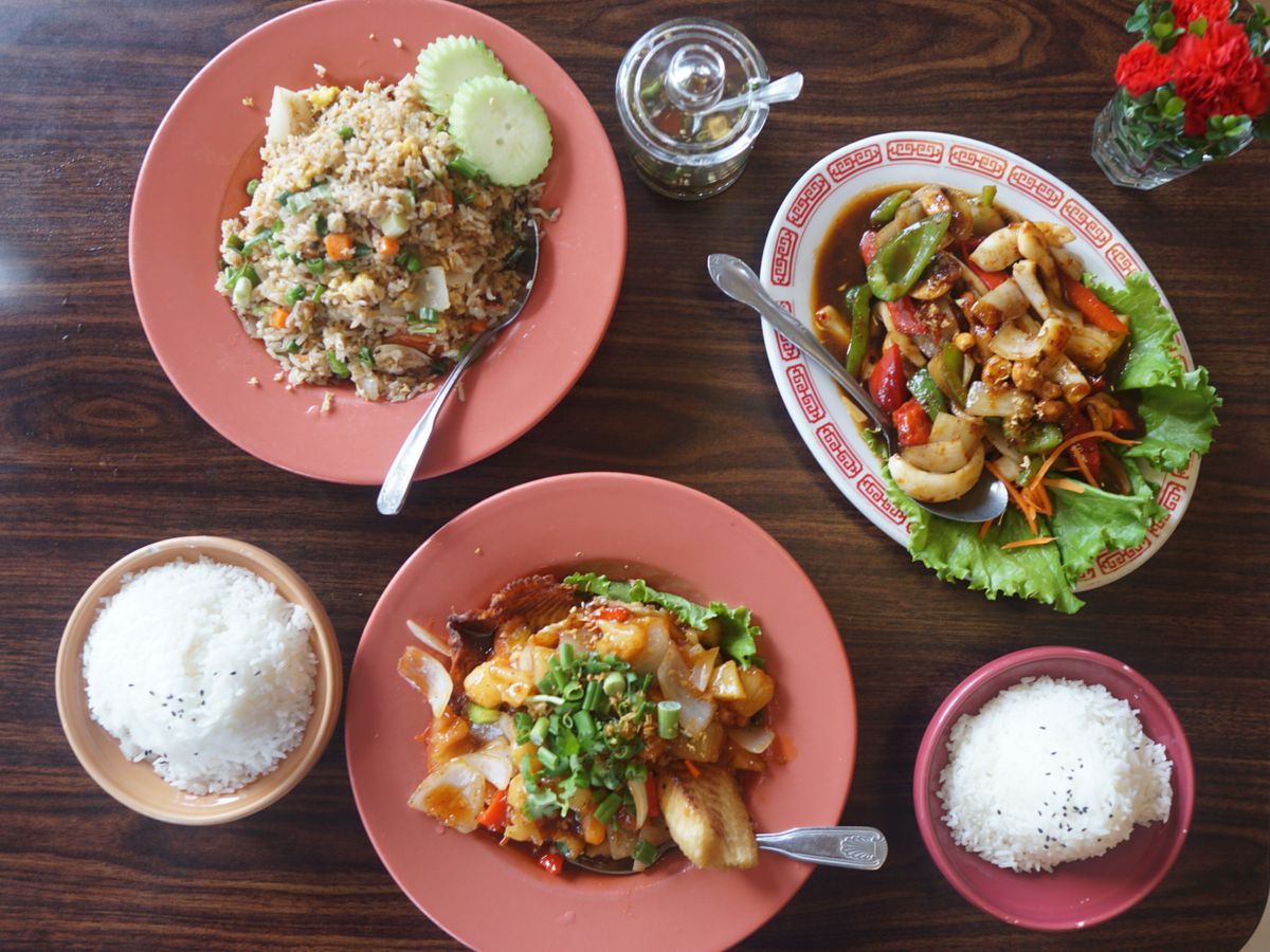 An overhead picture of three dishes at Thai Chili Jam: Crab fried rice, served with two pieces of cucumber, fried tilapia on a bed of cilantro, and a bunch of squid in chili jam. The three dishes are served on pink plates, with rice and jars of nam prik noom.