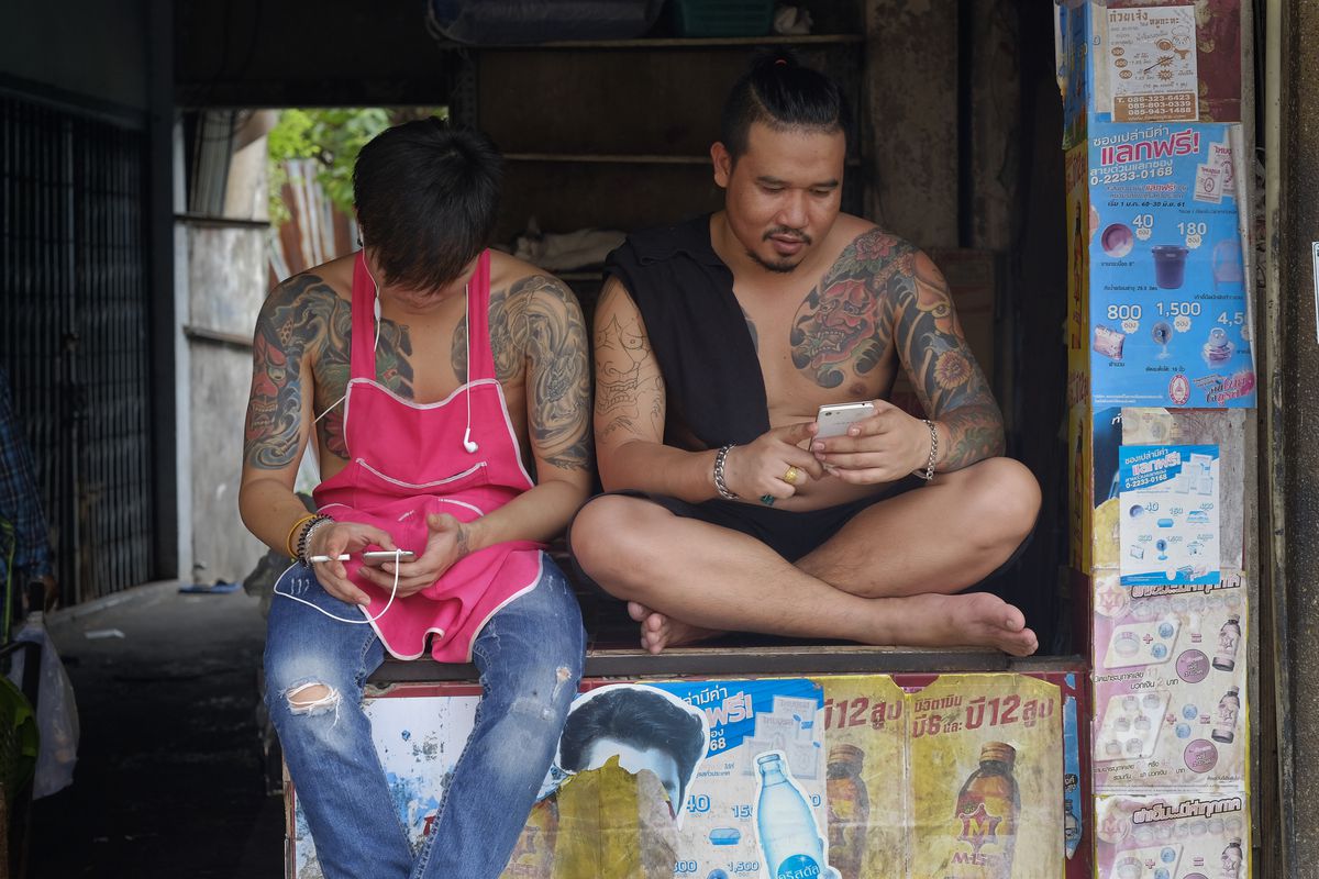 Two heavily tattooed Thai men sit in the back of a truck, each looking at his phone.