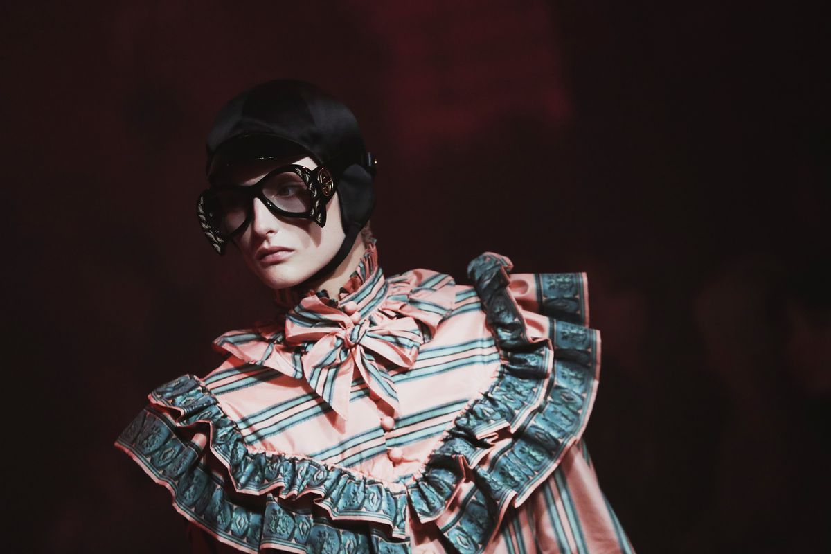 A model wears a blouse with a massive ruffle at the bust and neck, with oversized glasses and a bowl cut.