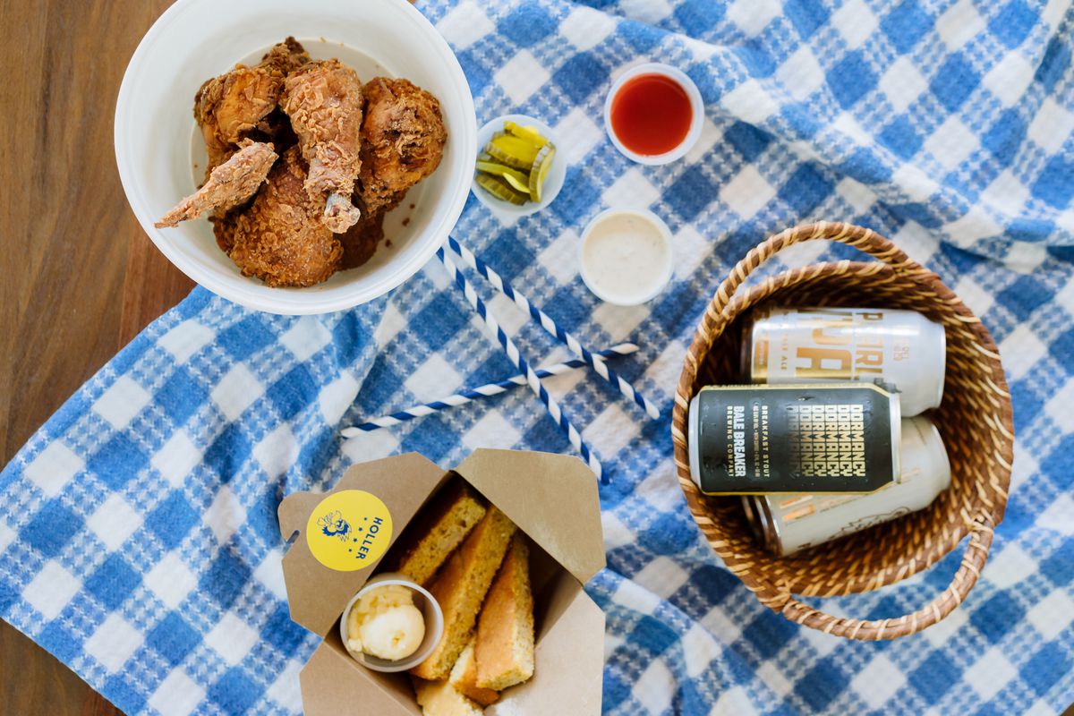 A bowl of fried chicken from Holler sits on a blue-and-white-checkered tablecloth, next to sticks of cornbread and a wicker basket full of canned beer