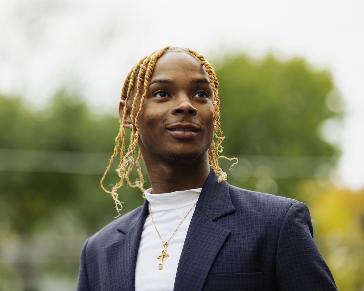 Crosby, in a navy blue suit, white turtleneck, and with a medium sized gold crucifix around his neck, looks up and into the distance, smiling. His bleached shoulder length braids move slightly in the wind; behind him, blurred, are green and yellow trees.