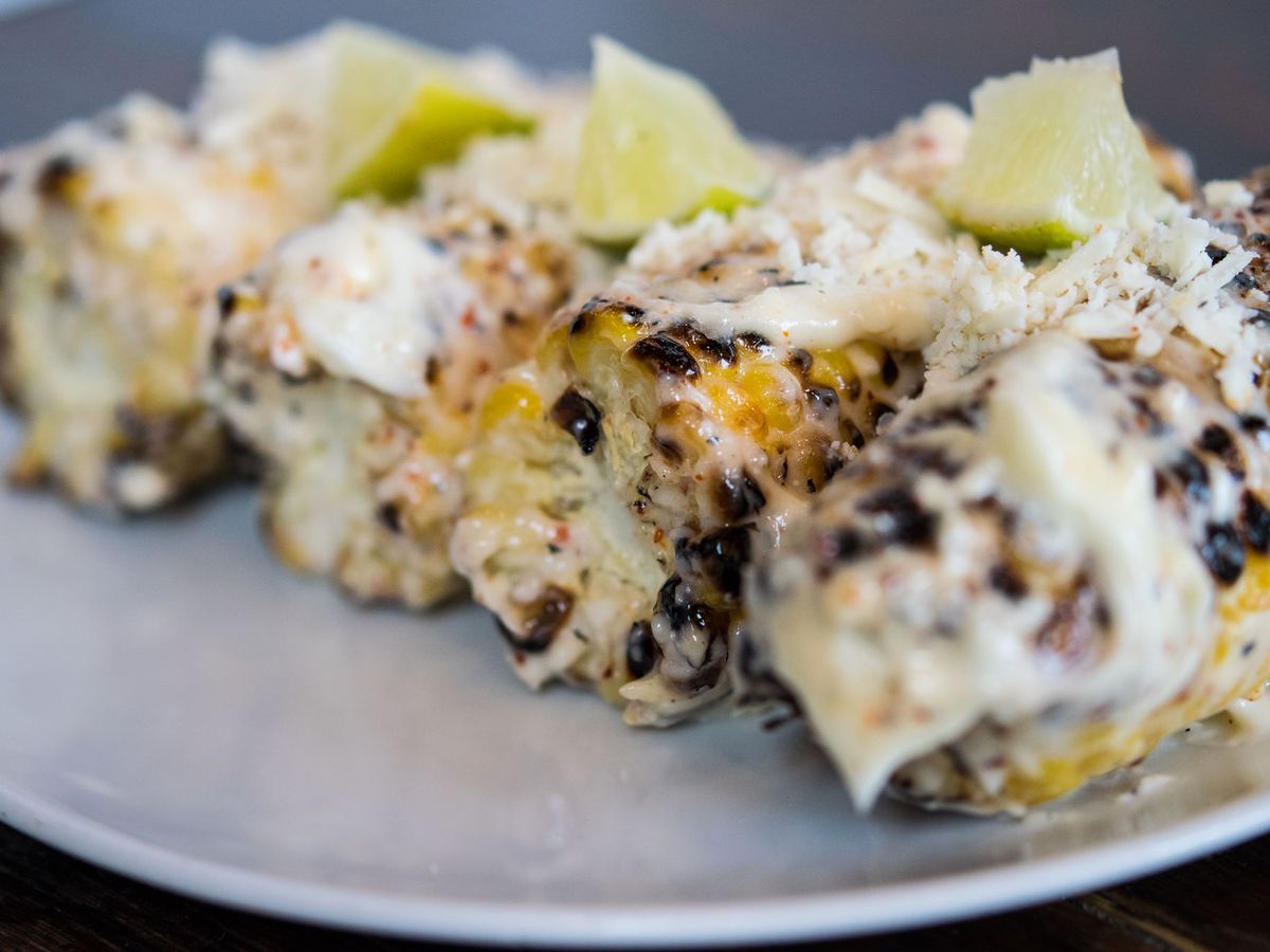 Four ears of corn, charred and topped with aioli, cotija, and lime wedges, sit side-by-side on a plate