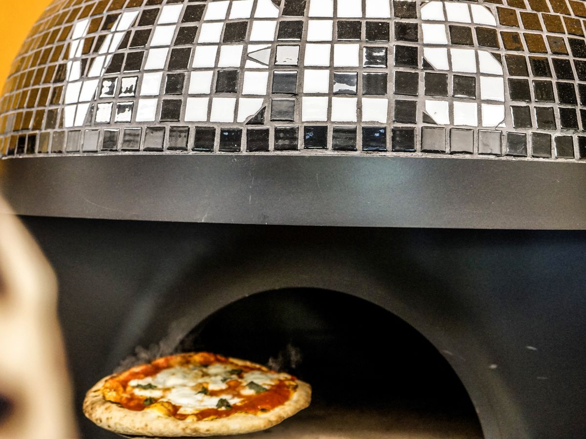 Black tiled dome of the wood-fire pizza oven at Holmes Slice with white tiles spelling out “Vibes”, a margherita pizza is coming out of the oven