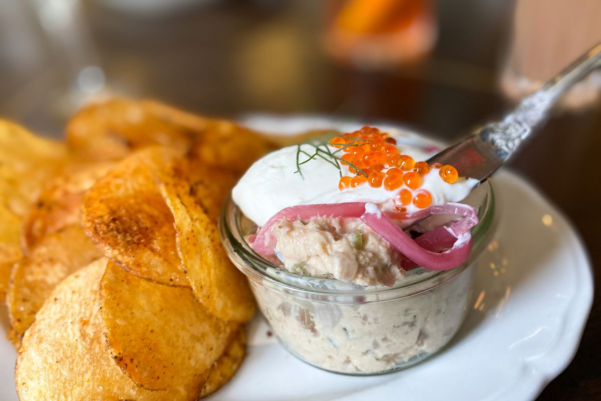 A white plate holds homemade potato chips and a glass ramekin with fish dip, topped with cream and caviar, with a silver spoon in it.