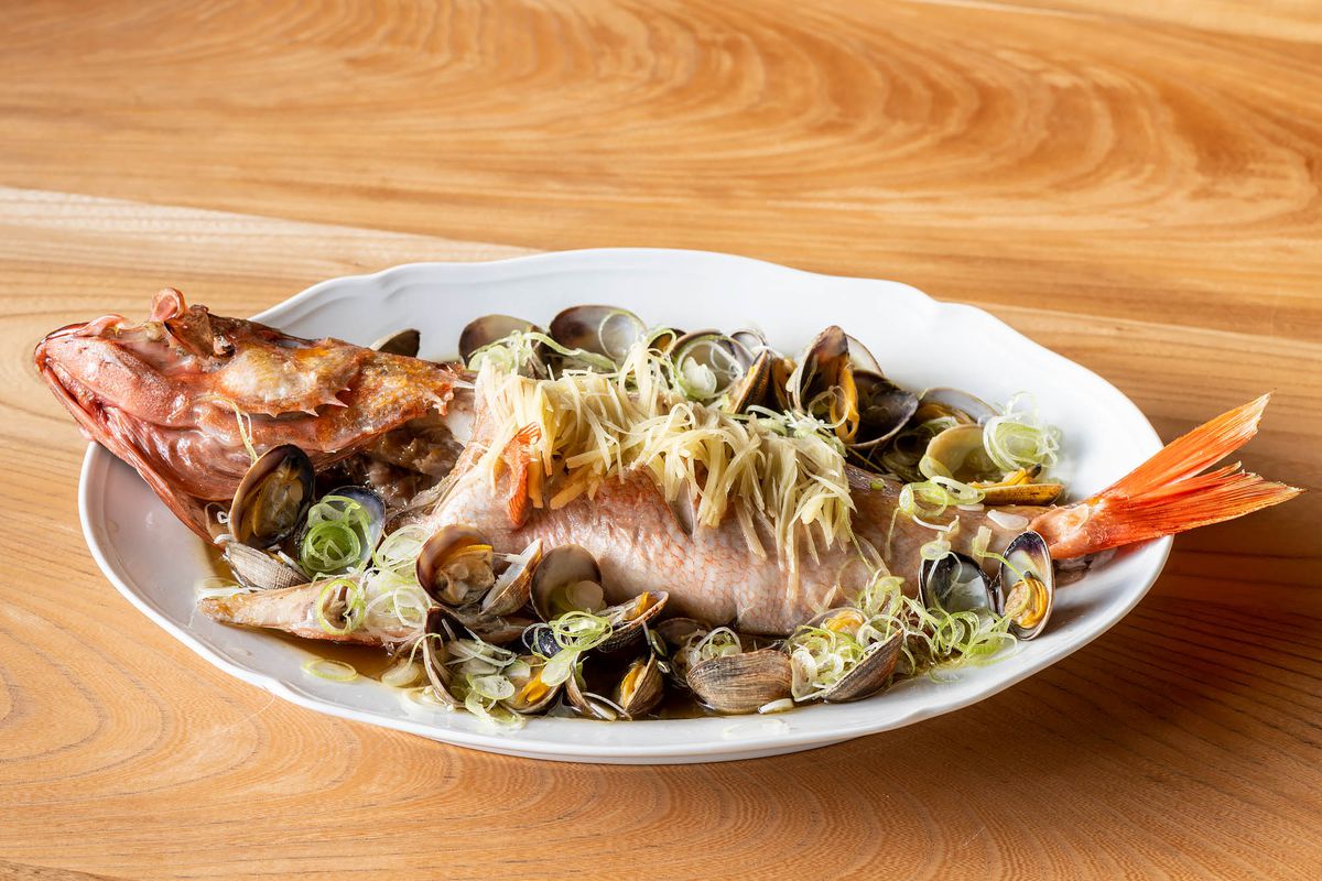 A steamed pink fish with clams and noodles served on a white plate against a light wood background.