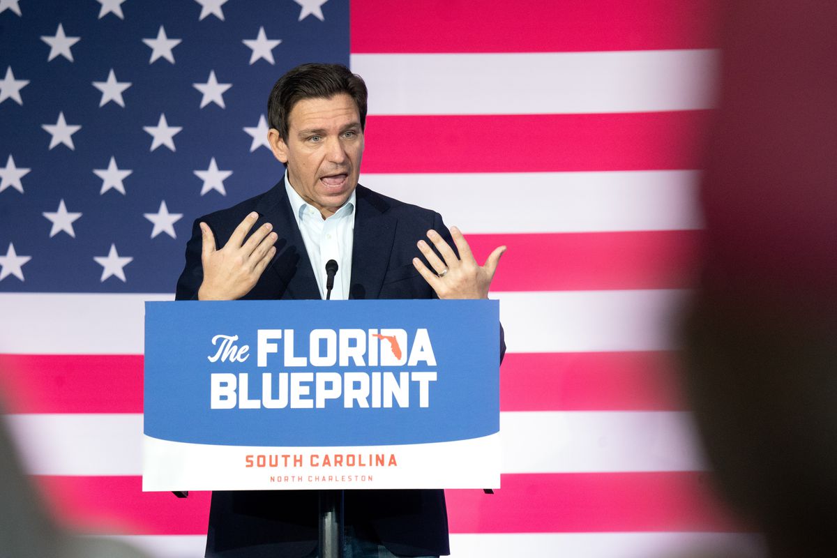 Florida Governor Ron DeSantis speaking from behind a lectern that reads “The Florida blueprint” while standing in front of a wall-sized American flag.