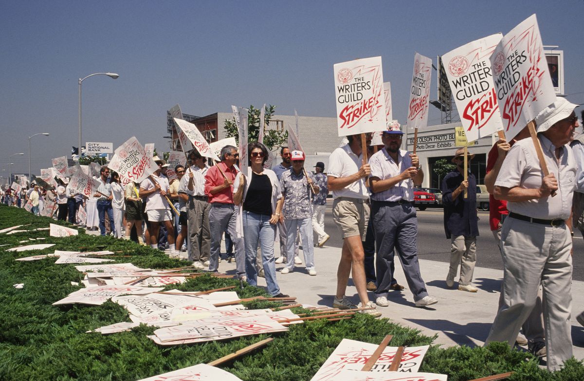 Against green grass and blue sky, a line of picketing writers hold signs identifying them as the 1988 striking Writers Guild. Behind them is a busy street with a bus stop and cars.