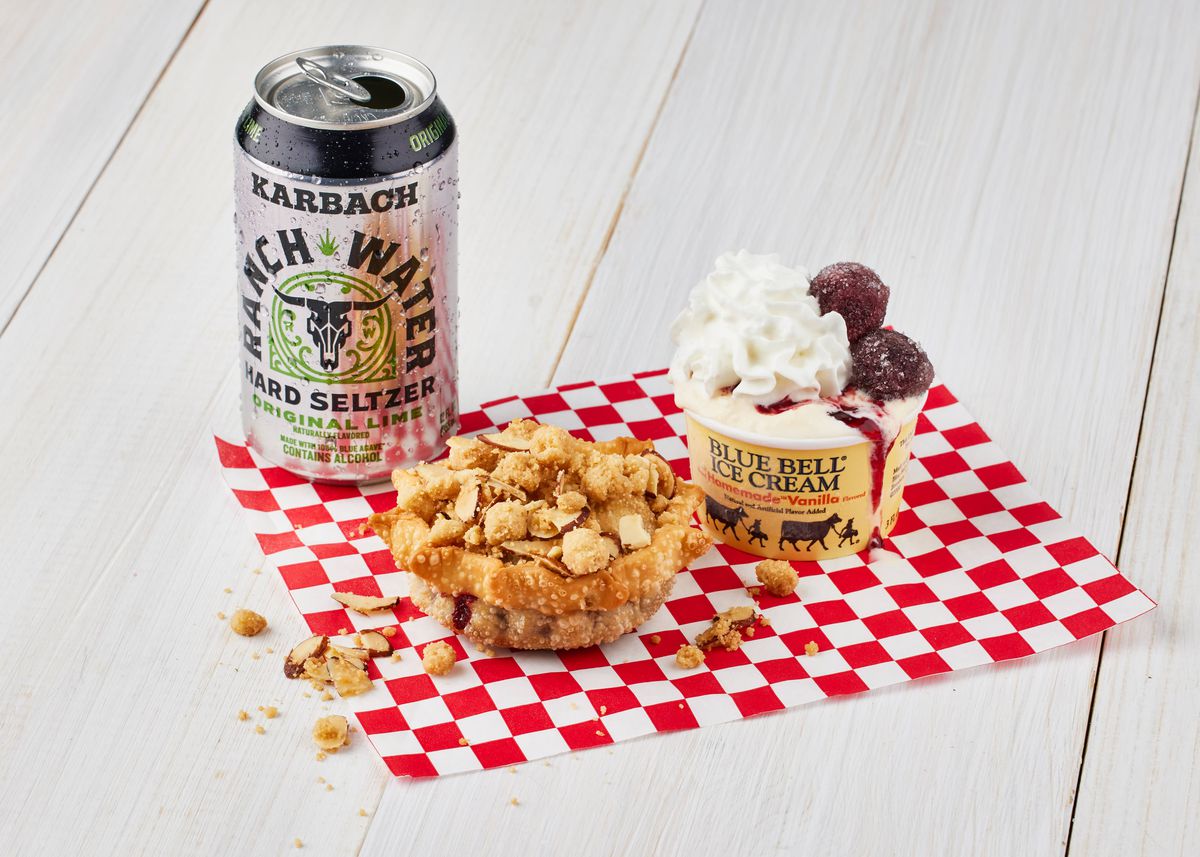On a red and white checkered napkin, a deep fried, hand sized cherry pie sits next to an individual sized Blue Bell vanilla ice cream topped with whipped cream and cherries. A can of beer sits to the left.