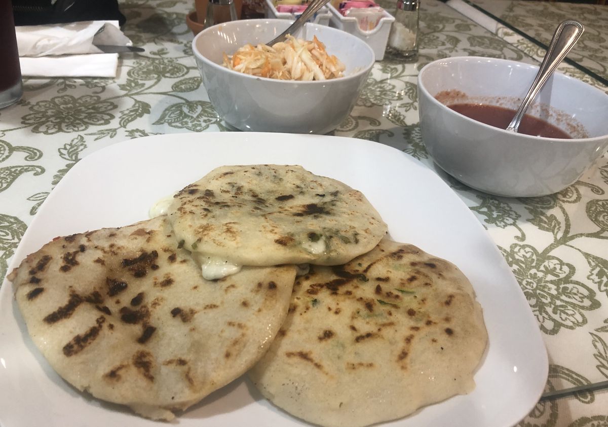 Three pupusas laid on a white plate with two white bowls filled with pickled cabbage and red tomato sauce sit on a green and white patterned table.