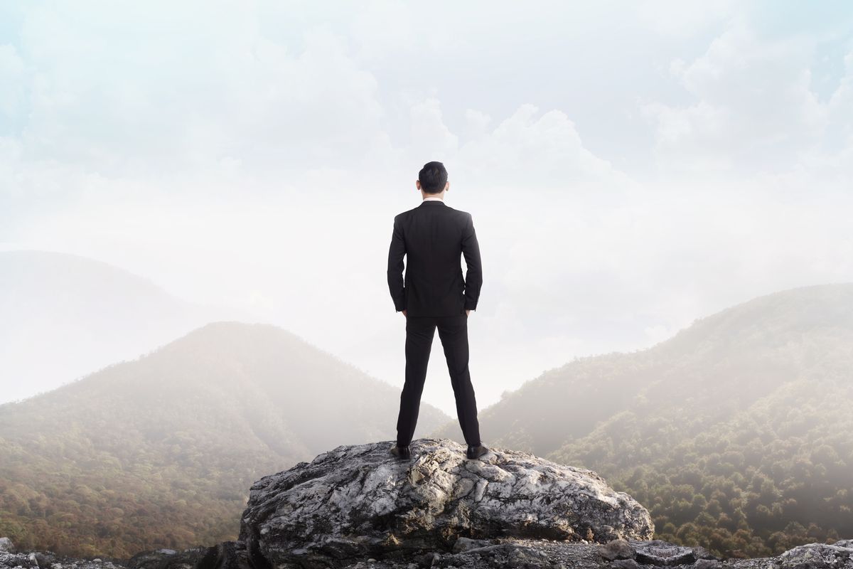 A stock image of a man in a business suit, hands in pockets, standing on a cliff and looking at the mountains in the distance.