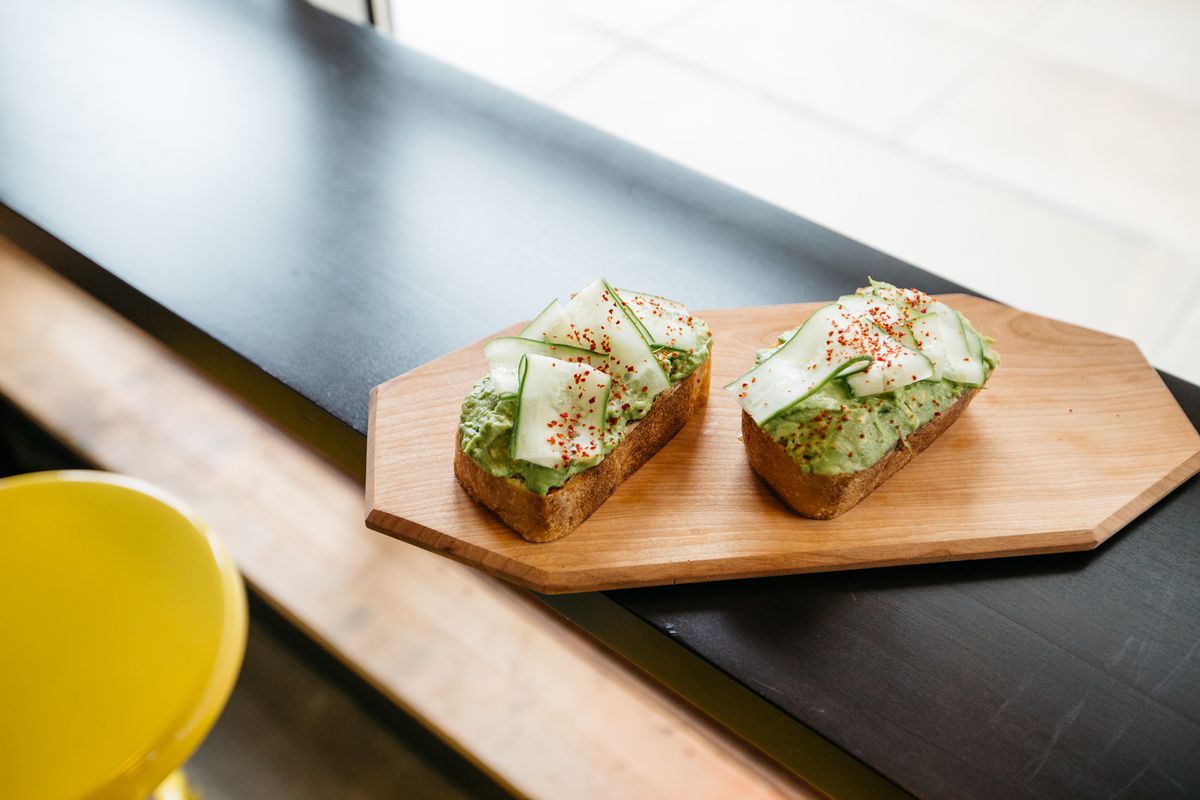 Two slices of avocado toast with cucumber and a sprinkle of red spice sit on a wooden board in a window. 