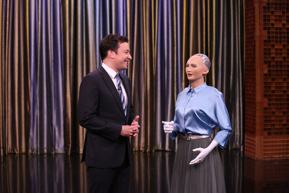 Man in suit and robot woman in skirt and button up in front of curtain.