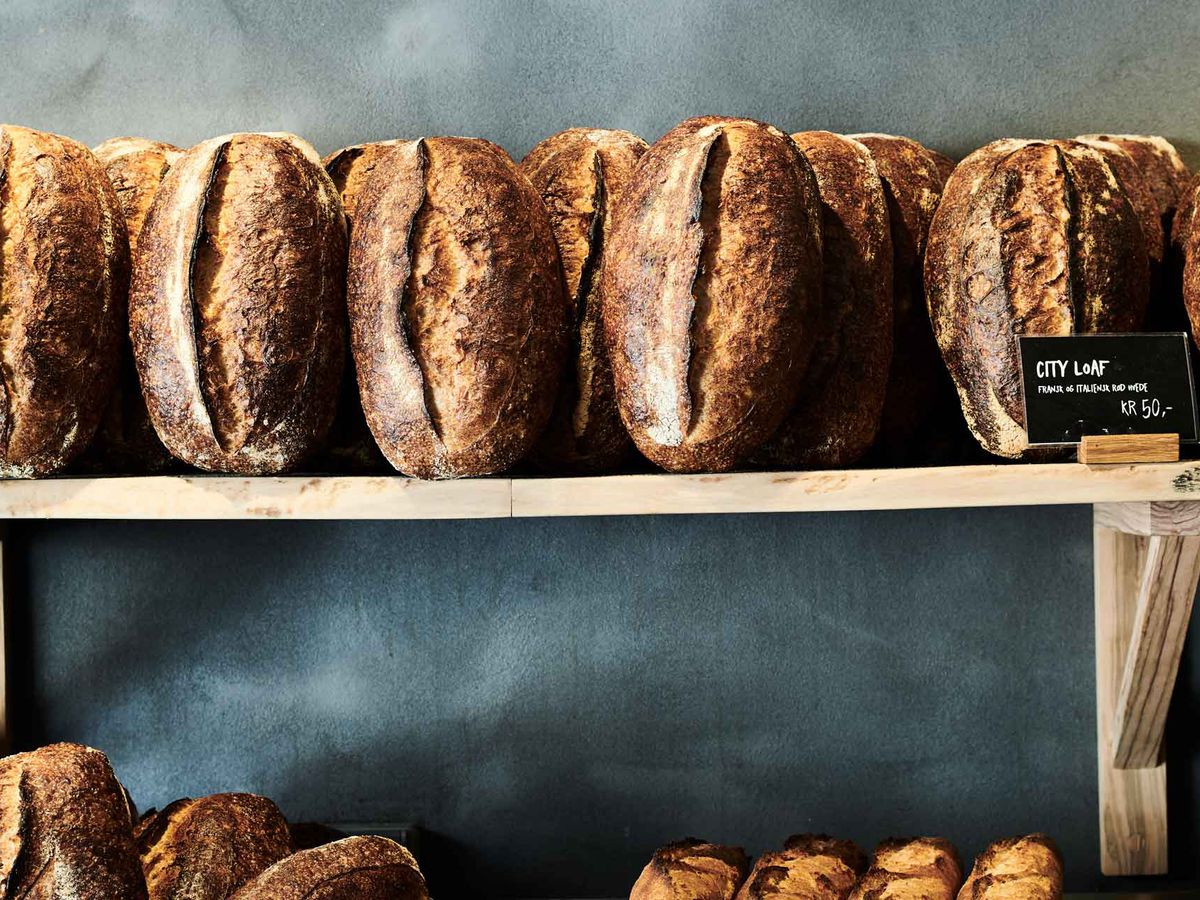 A wooden shelf holds loaves of sourdough bread rested on end against a slate wall behind, with a small chalkboard sign that reads “City Loaf”.