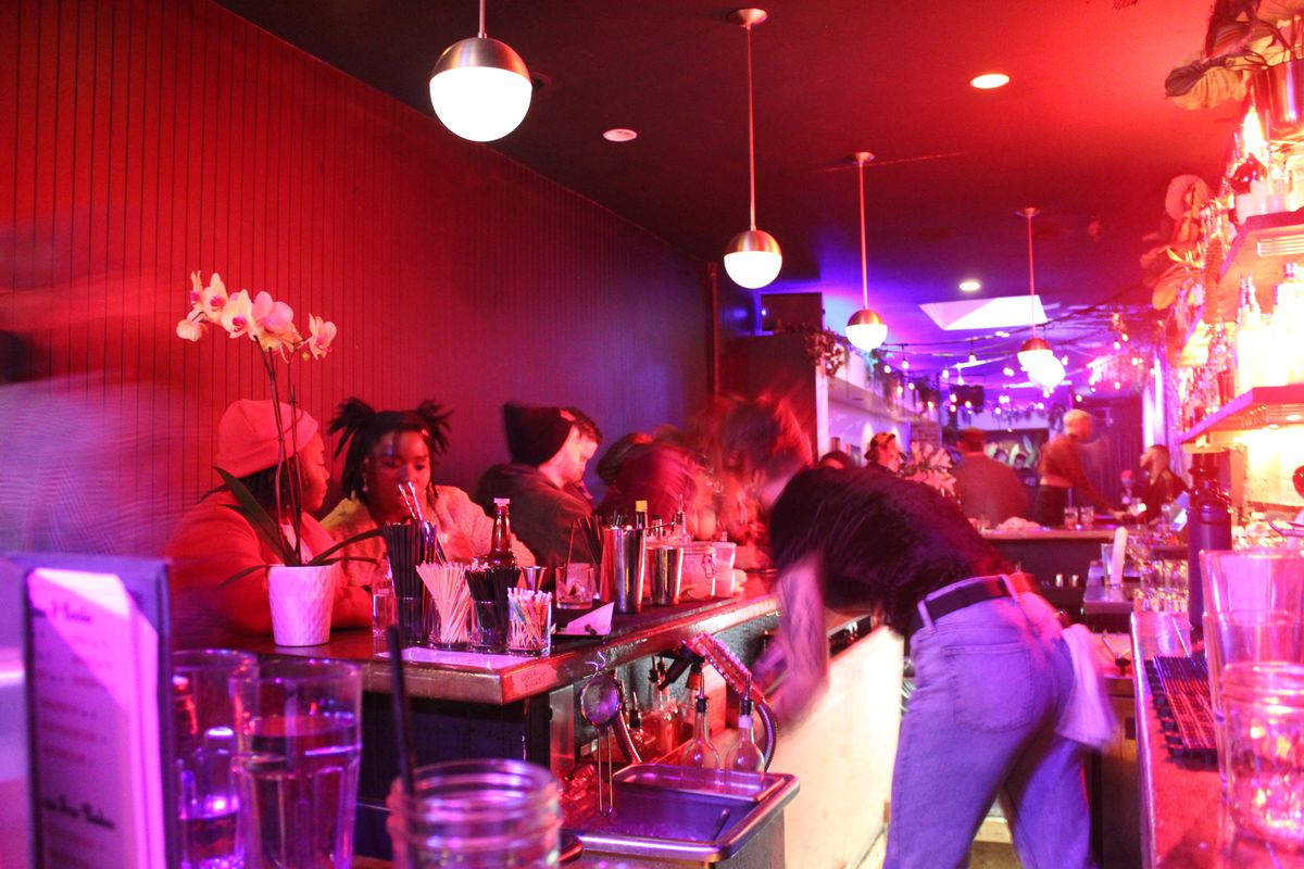 A pink-and-red-hued bar space at Rebel Rebel is blurry with nightlife, while a bartender bends over to make a drink.