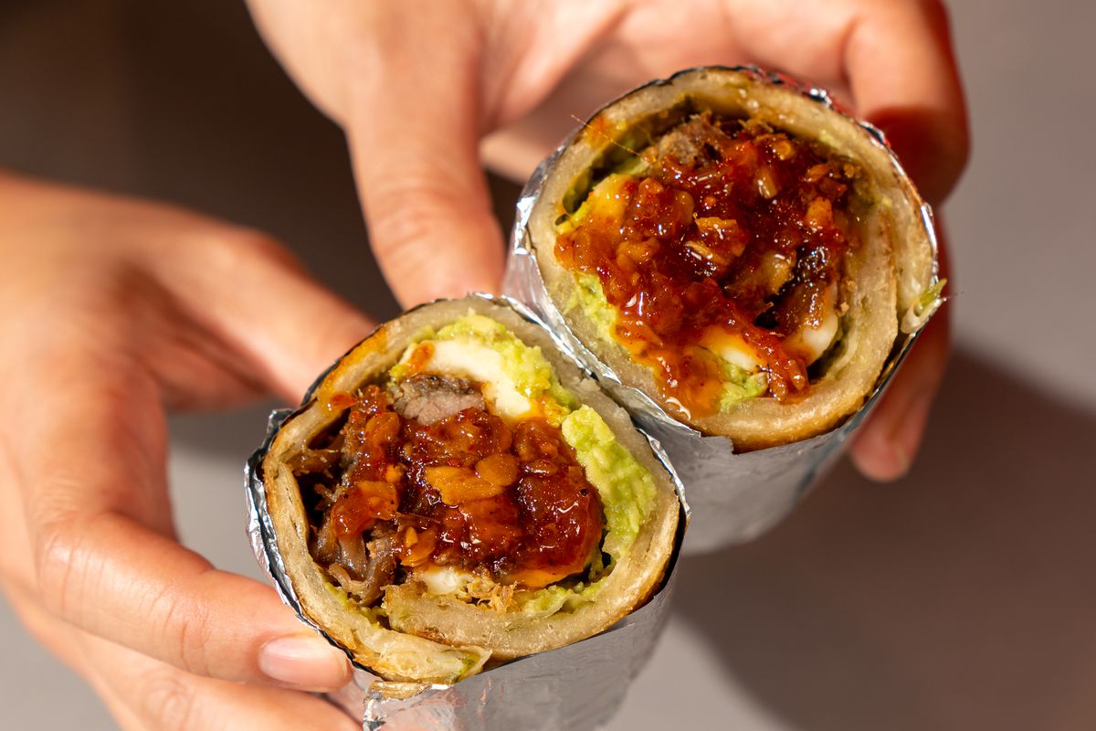 Hands hold up a burrito made with scallion pancakes, that’s been cut in half and wrapped in tinfoil.