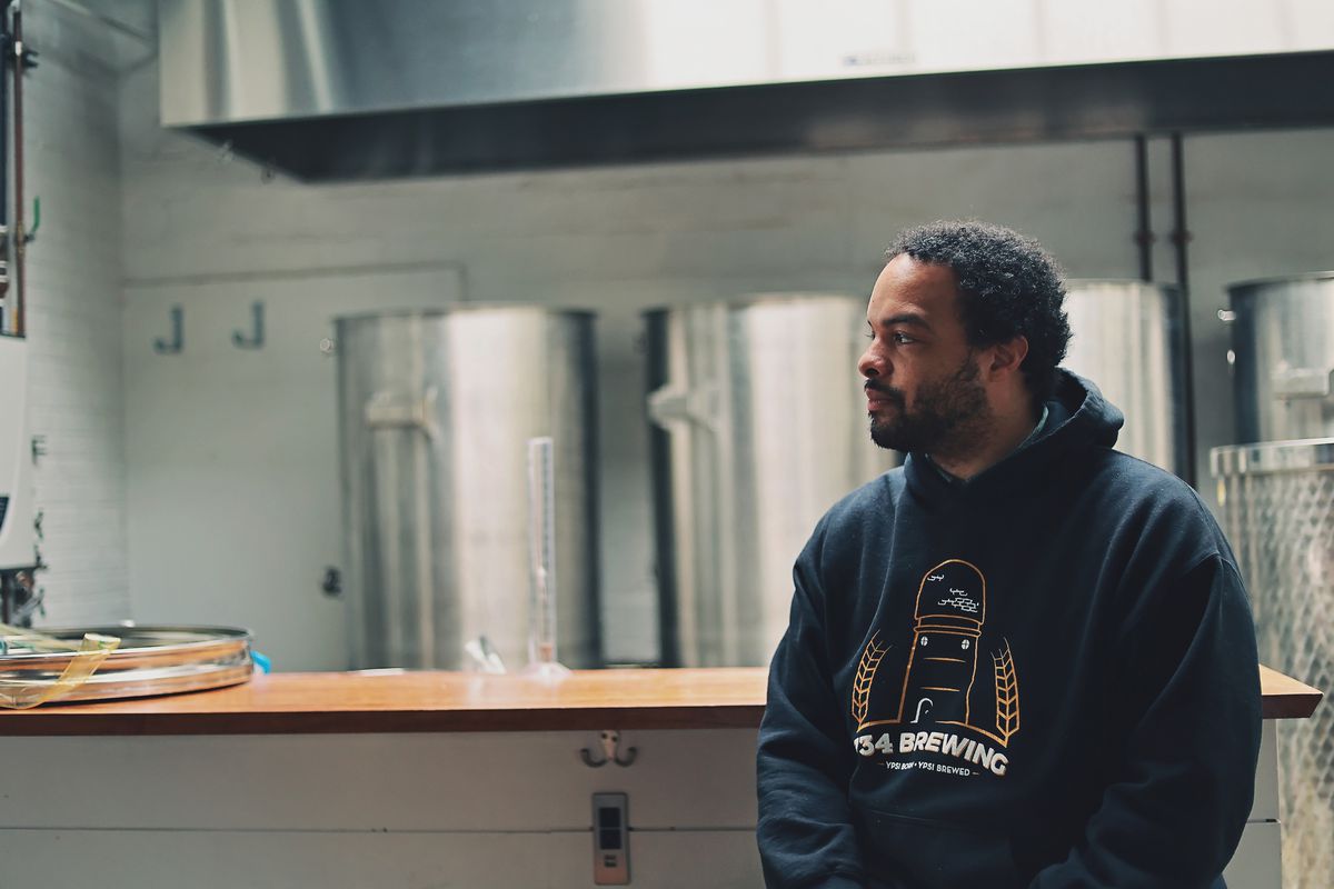 Brian Jones-Chance stares to the left in his black hooded 734 Brewing sweatshirt in front of beer brewing equipment at 734 in Ypsilanti.