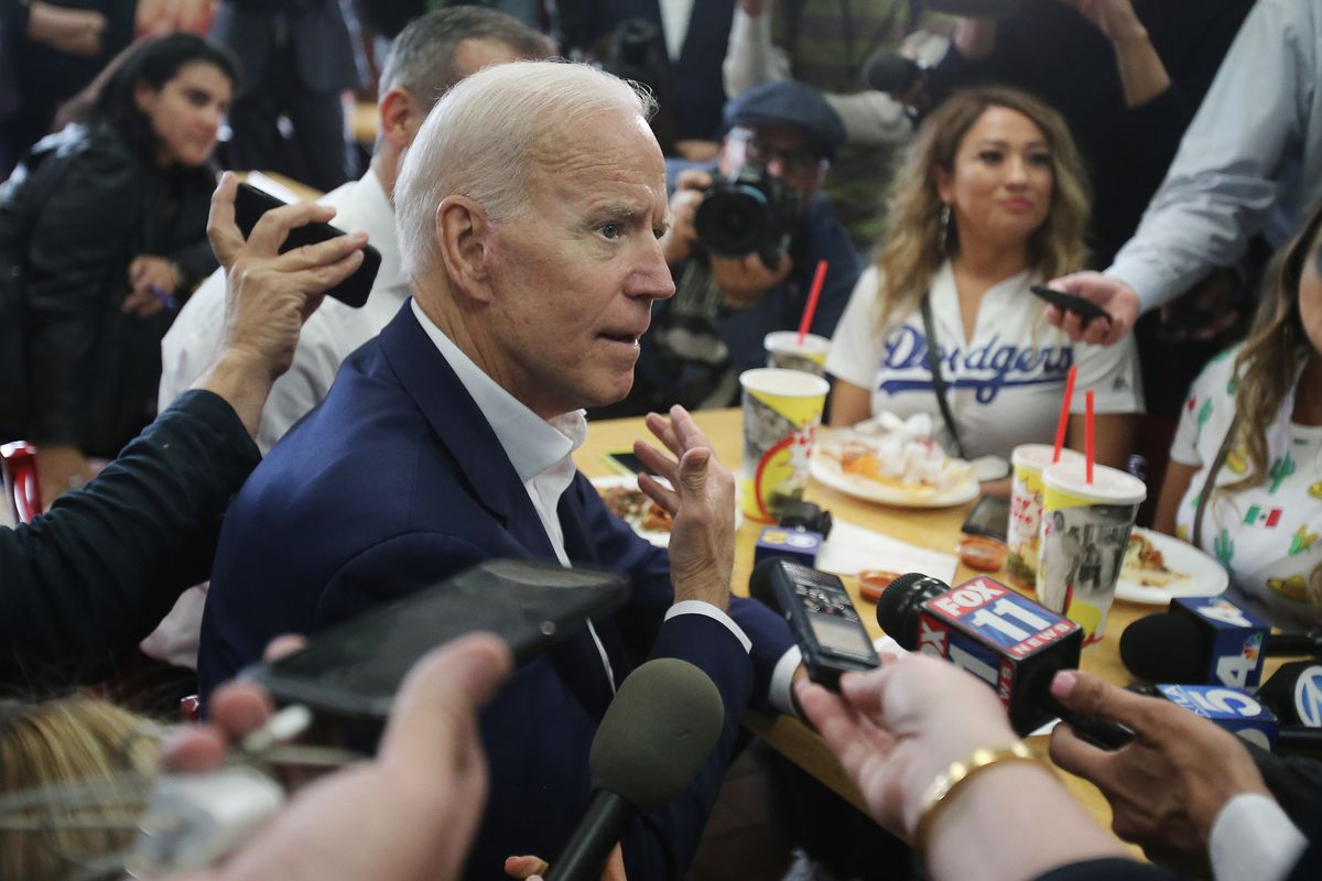 joe biden seated at a table, surrounded by reporters’ hands