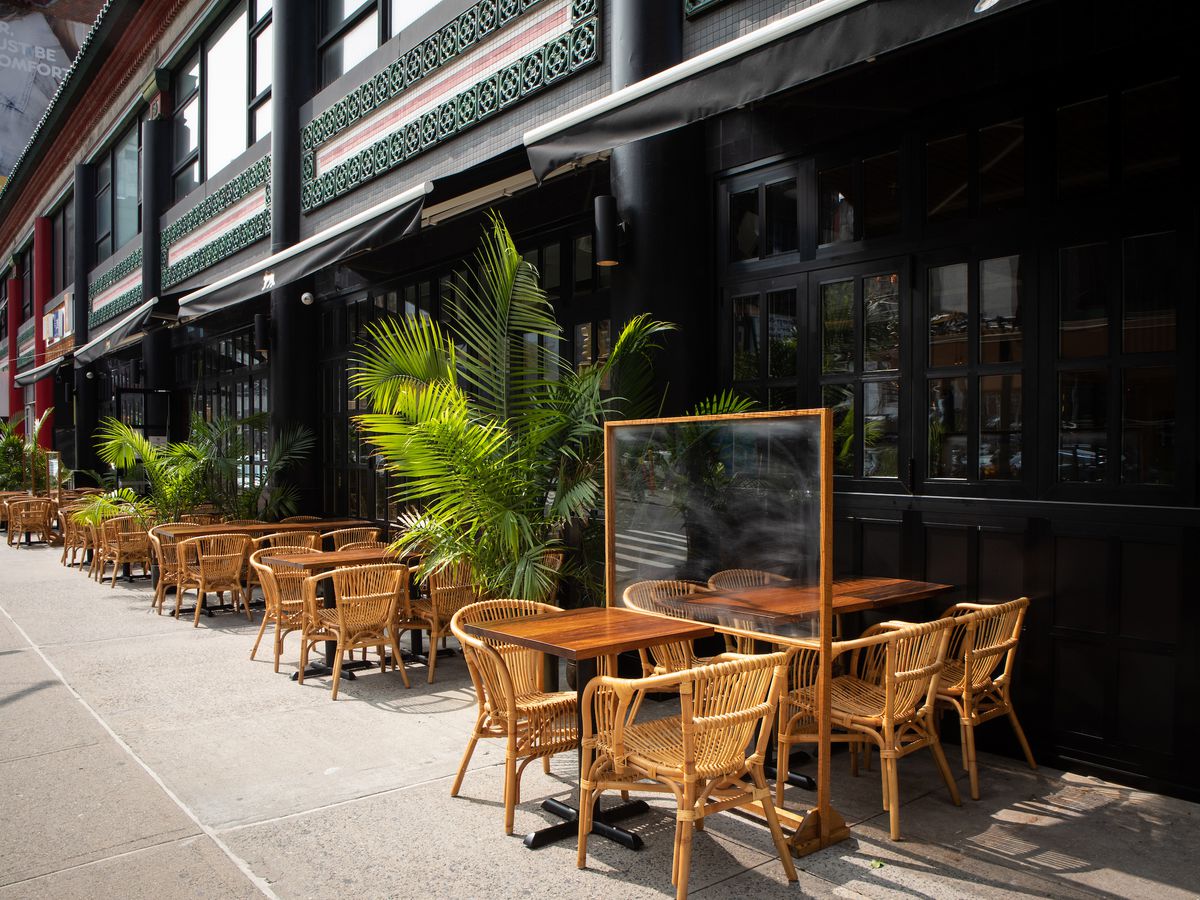 Light wicker chairs and wooden tables with green palm plants in between set up on the sidewalk alongside the exterior of the restaurant