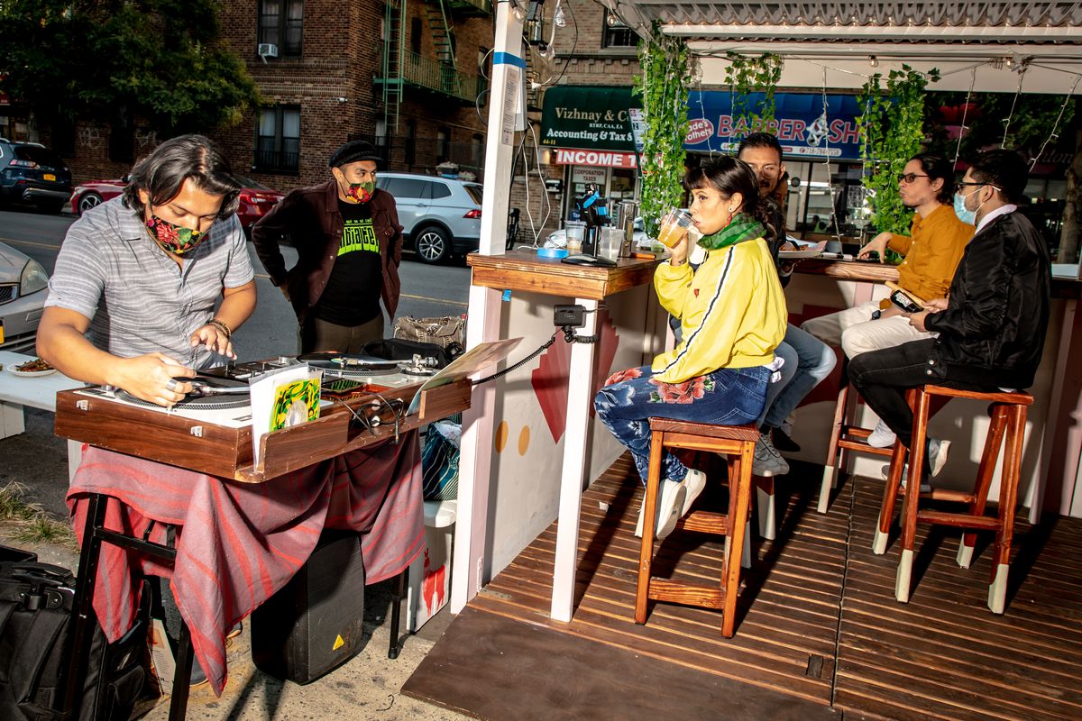 Six people in an outdoor seating area. Four sit on stools, one wearing a mask. One man in a mask plays records at a DJ stand. One man in a mask stands by him.