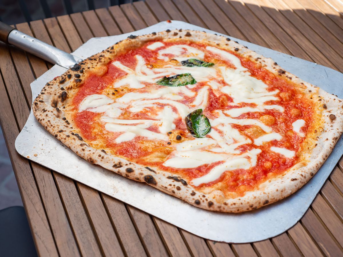 A whole wood-fired pizza resting on a steel pizza paddle that is set on a wooden table