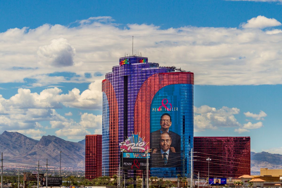 A hotel with blue, red, and purple windows with mountains and a blue cloudy sky behind it.