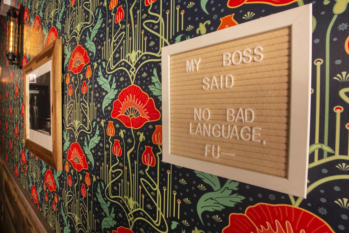 Colorful floral wallpaper lines a wall with a sign that reads “My boss said no bad language. Fu—.”