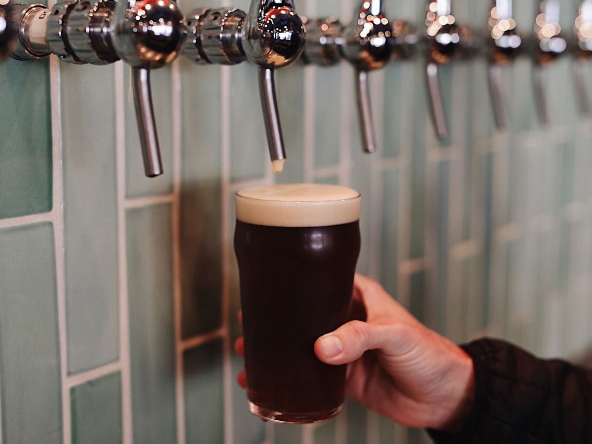 A dark beer being poured from a silver tap in front of light green teal tiles.