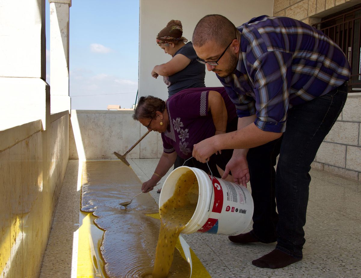 A man pours a yellow mixture from a large bucket onto a long plastic sheet, set in an outdoor covered patio. Two women stand nearby spreading the mixture with broom-like tools.