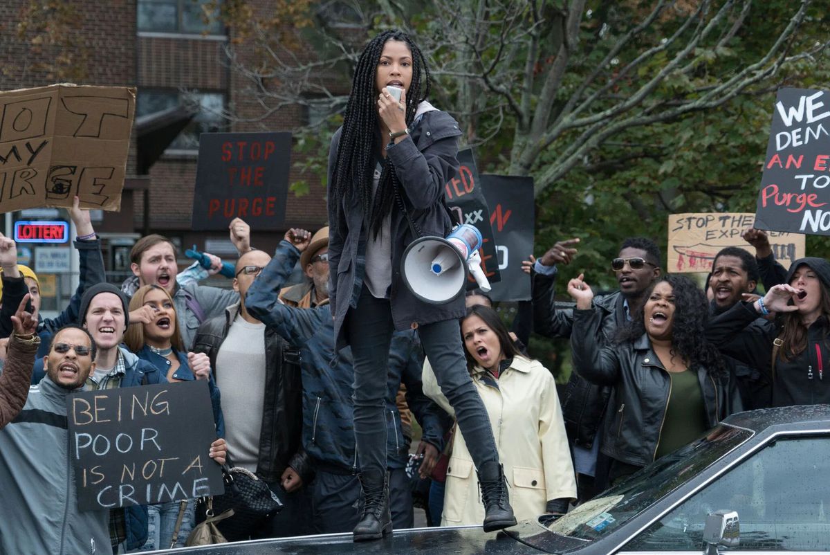 A young woman standing on top of the hood of a car in front of a group of protestors in The First Purge.