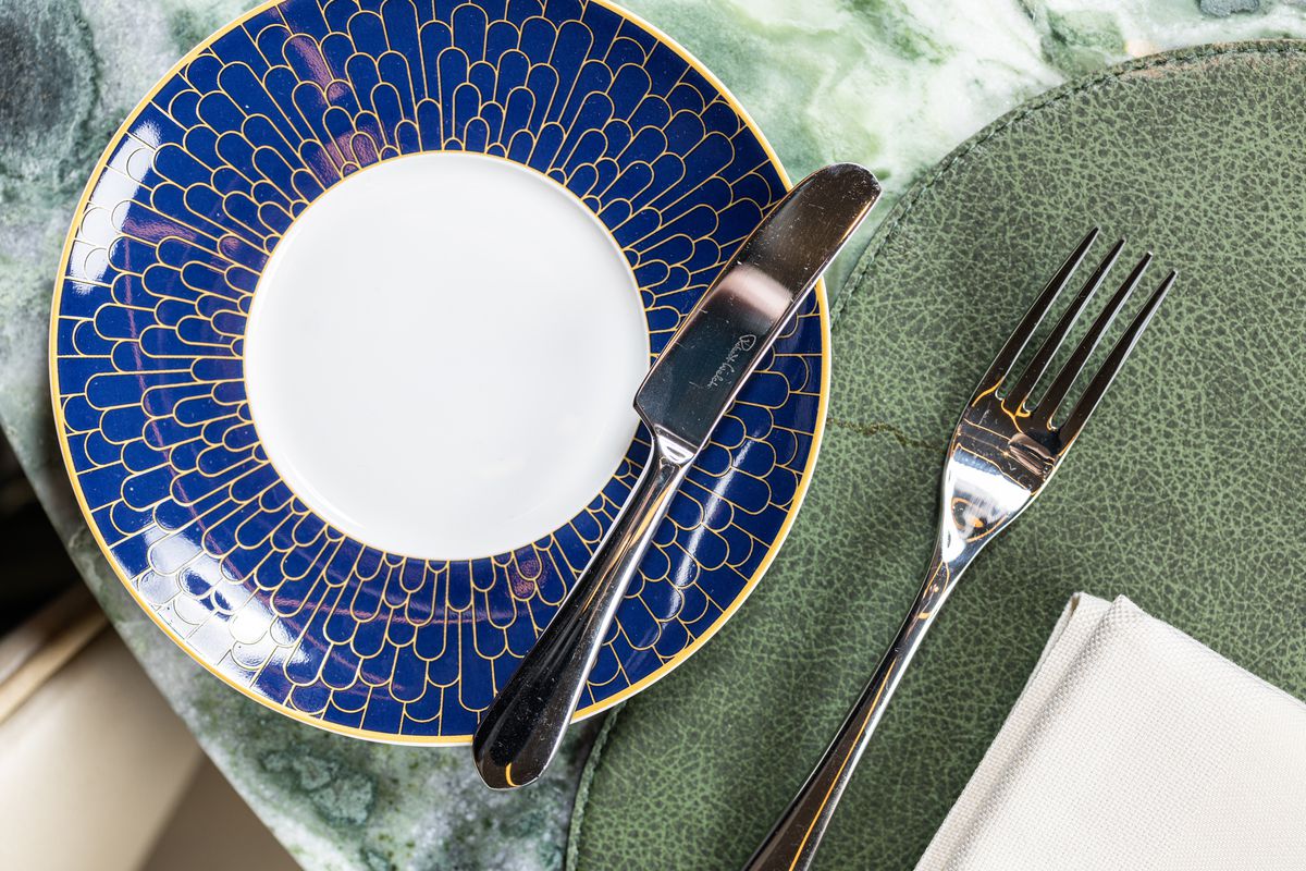 A blue plate with gold flourishes and a white center sits on a marble table top, garnished with silverware.