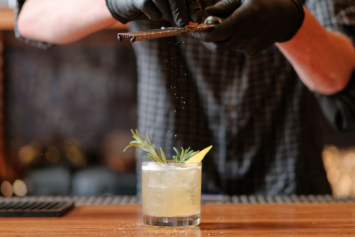 Two gloved hands using a microplane to grate ginger over a high-ball glass with rosemary sticking out.