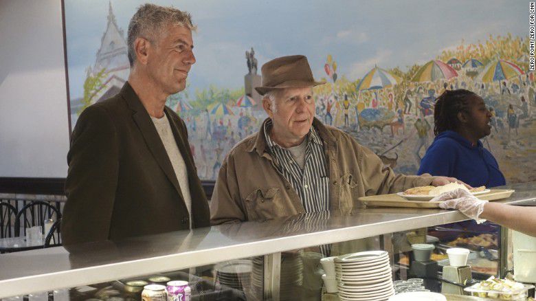 Two white men, one tall, the other short and wearing a fedora, stand in front of a cafeteria counter.