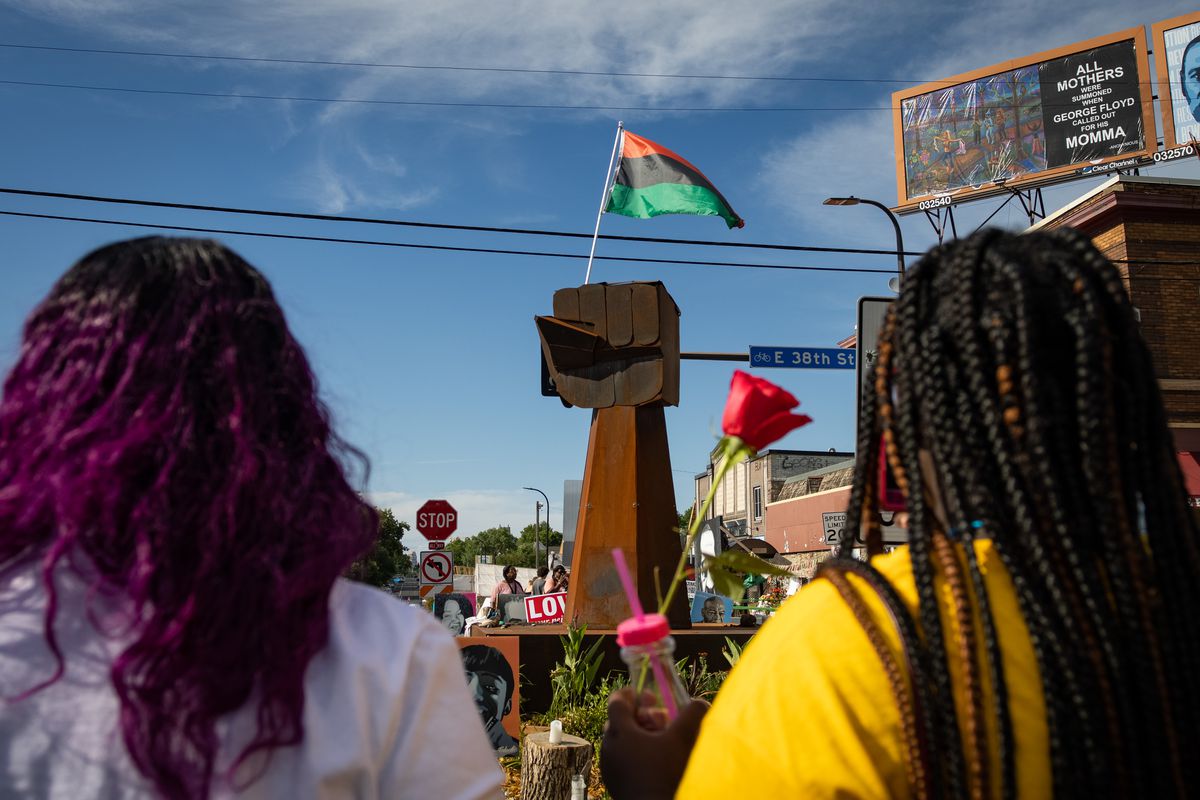 Two people, one with long black braids wearing a yellow shirt, and one with black and purple curls wearing a lilac shirt, stand with their back to the camera. They are facing a sculpture of a brown raised fist flying the Pan-African flag, which has a red, black, and green stripe. The sky is blue and a red rose is suspended between them. 