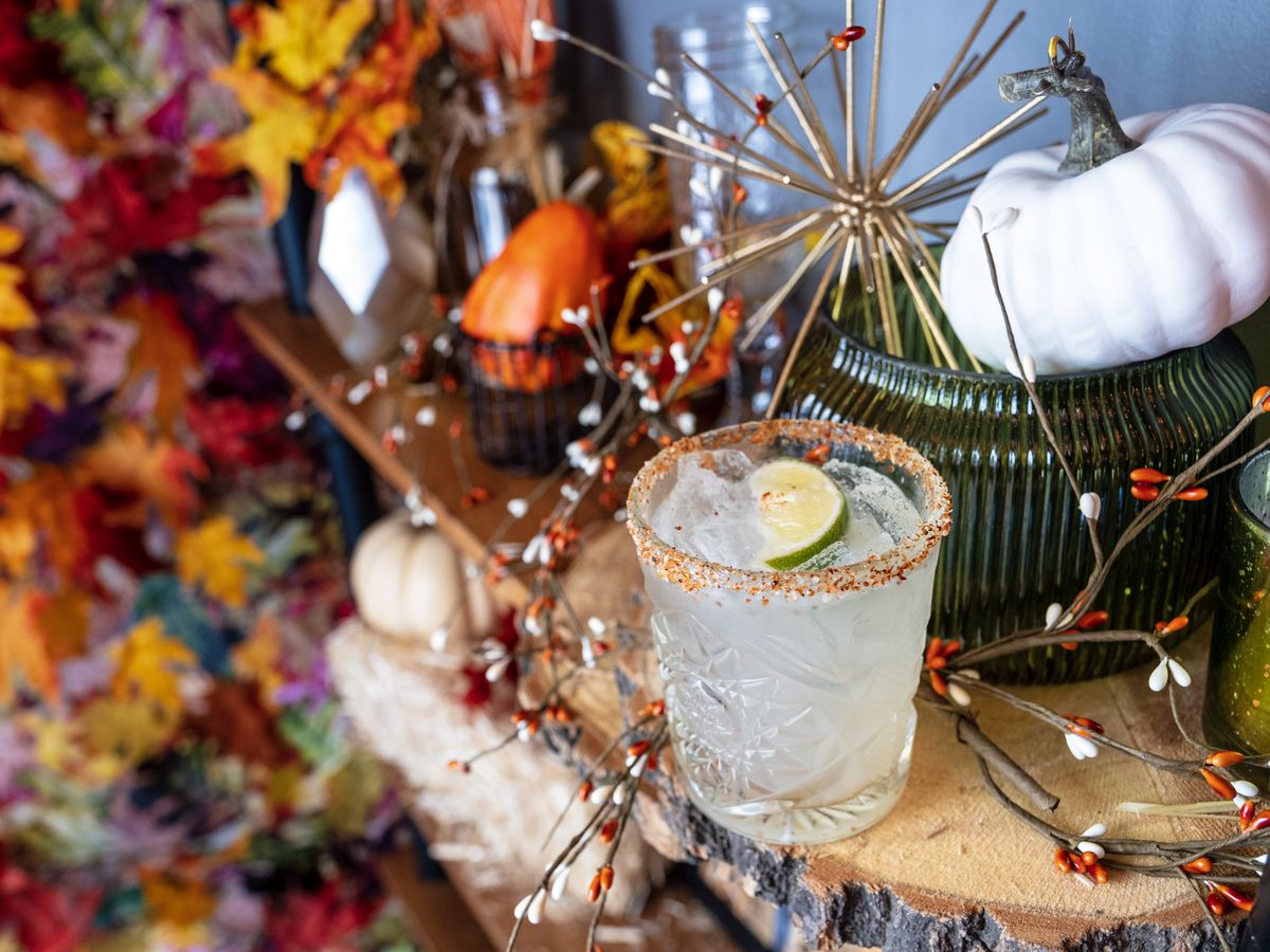 A cocktail in a short glass with a sugared rim and lime garnish on a table with a backdrop of colorful leaves and a round white gourd.