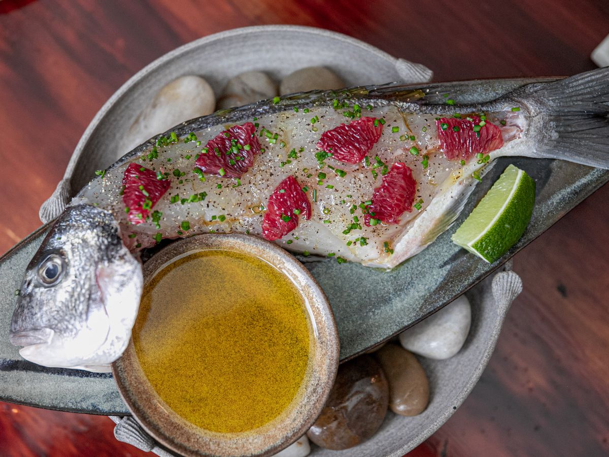 Whole fish, dotted with splotches of grapefruit and herbs, served with a bowl of sauce. 