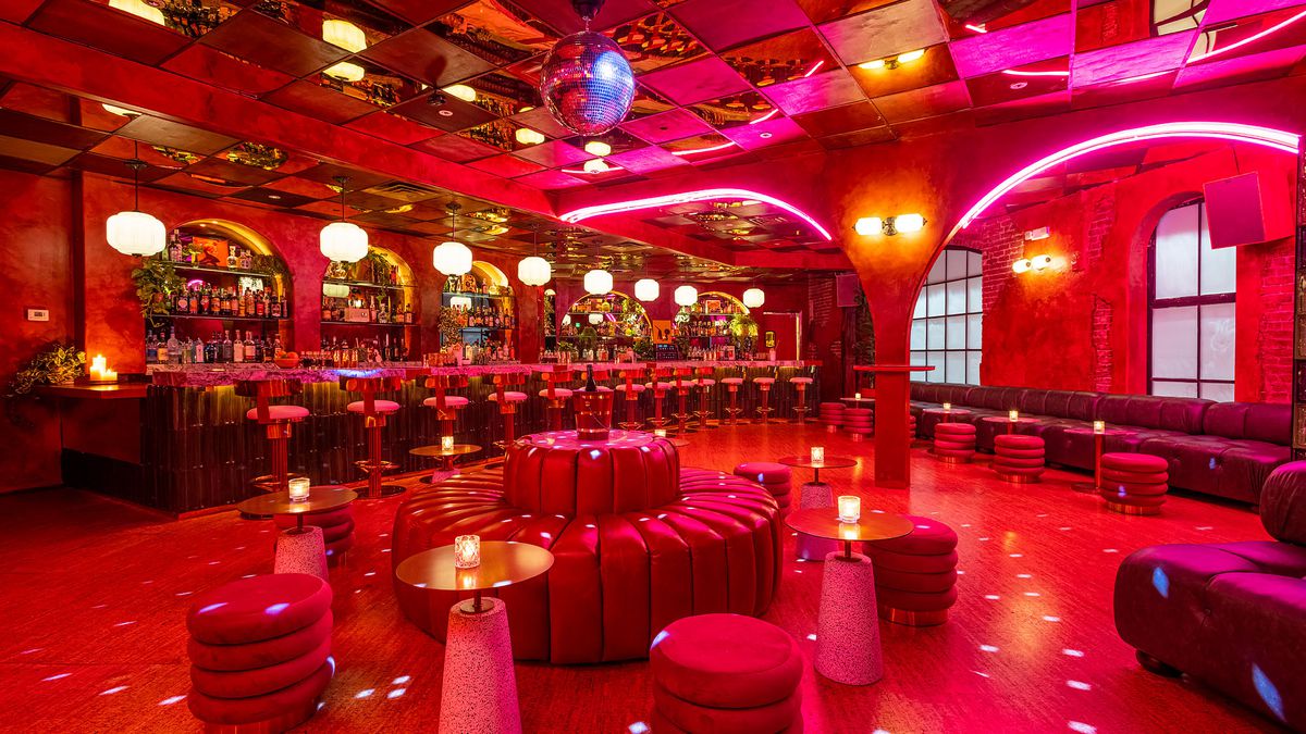 A disco ball shines specks of light across a red-hued room with a bar and round plush couches.