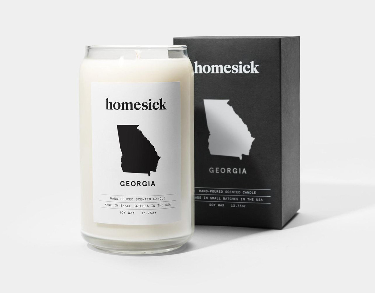 A candle and a box sit on a white surface. The candle and box have stickers with the silhouette of the state of Georgia and a word which reads: homesick.