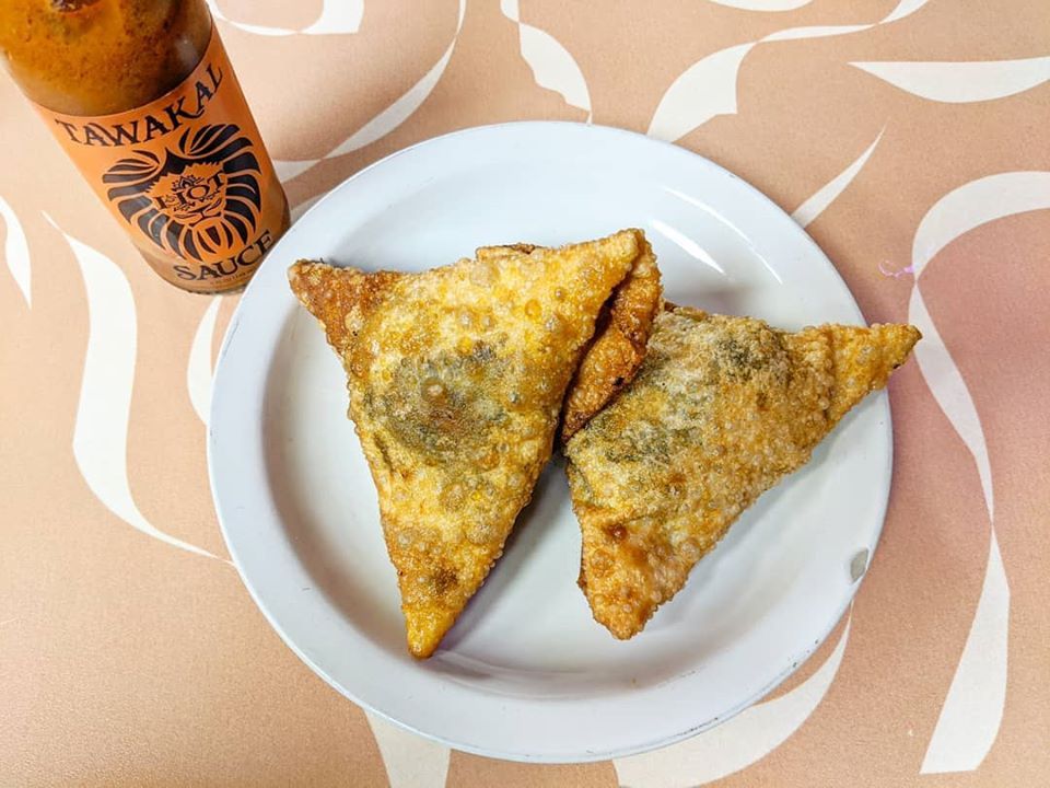 Fried samosa-style dumplings sit on a white plate on a peach and white table, with a bottle of orange hot sauce to the side