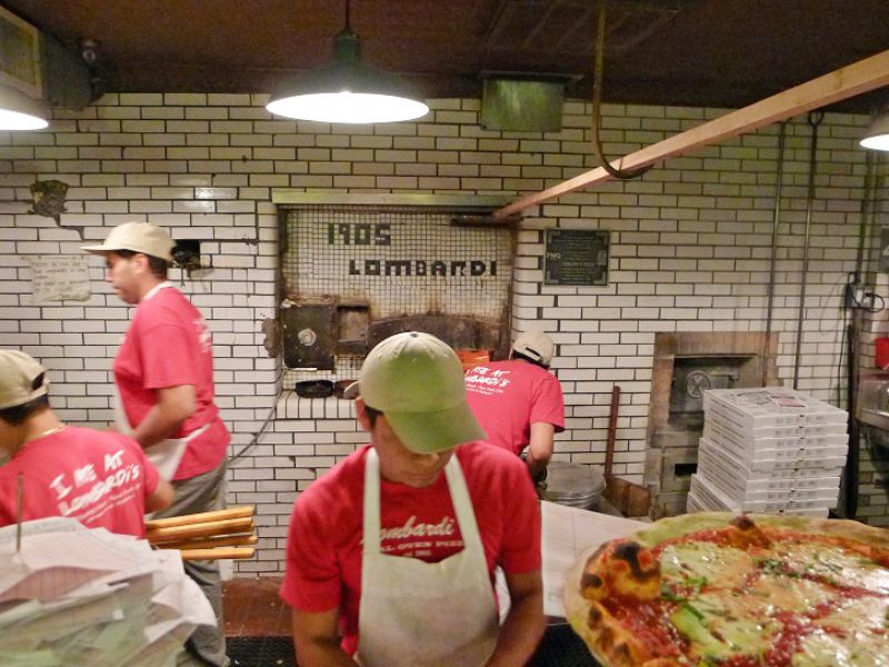 Four employees in red shirts and white aprons work in a kitchen, behind them the words “1905 Lombardi” are etched into a tile wall