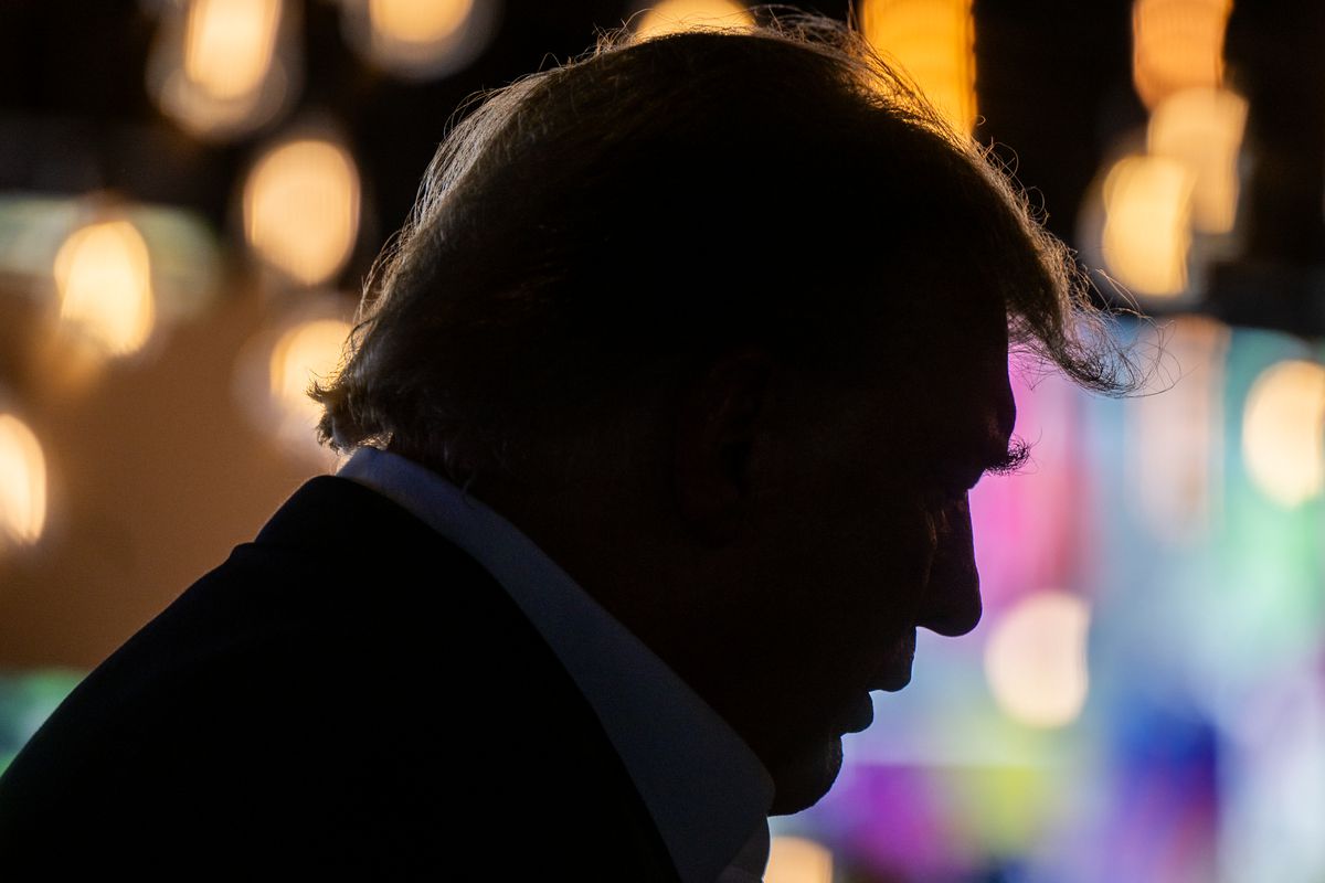 Trump’s face in silhouette, dark against blurry background lights.
